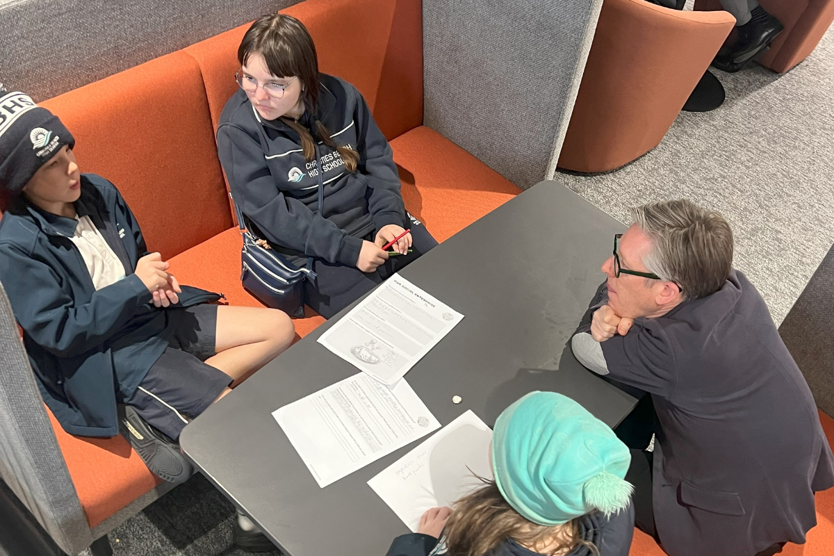 Students and an adult seated around a table, discussing worksheets in a quiet study space.